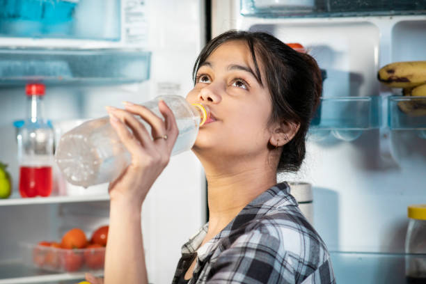 Woman drinking cold water while standing front of fridge in hot summer.