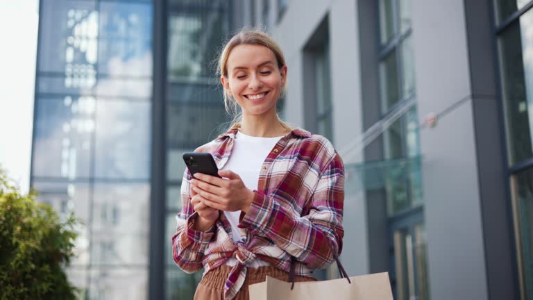 Happy excited young woman with packages receiving discount for shopping while checking email box on mobile tapping message receive cashbask looking at camera with perfect smile.