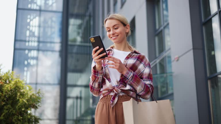 Happy excited young woman with packages receiving discount for shopping while checking email box on mobile tapping message receive cashbask sharing sales news walking in urban street shopping mall.