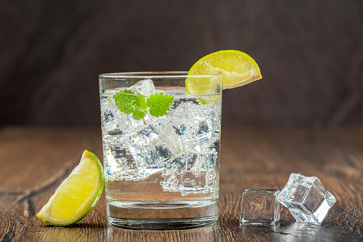 A glass of refreshing mineral water with ice cubes, mint leaves and lime slices on a wooden bar counter. Wooden background.