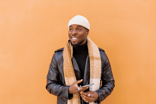 Cheerful bearded African American male wearing leather jacket scarf and hat standing against light brown wall and looking away with happy smile