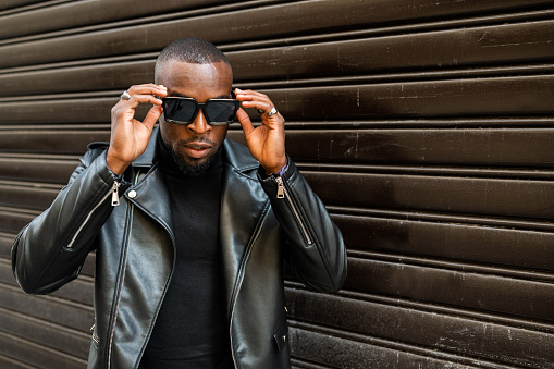 Serious masculine African American male in black leather jacket lowering stylish sunglasses and looking at camera with attitude while standing against brown wall