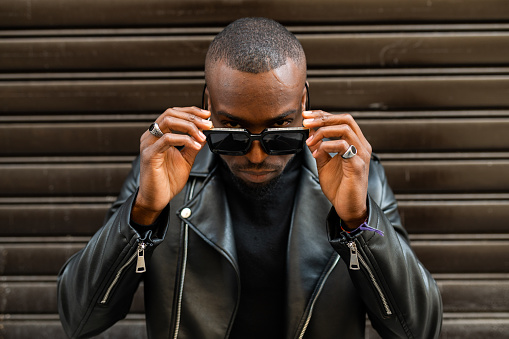 Serious masculine African American male in black leather jacket lowering stylish sunglasses and looking at camera with attitude while standing against brown wall