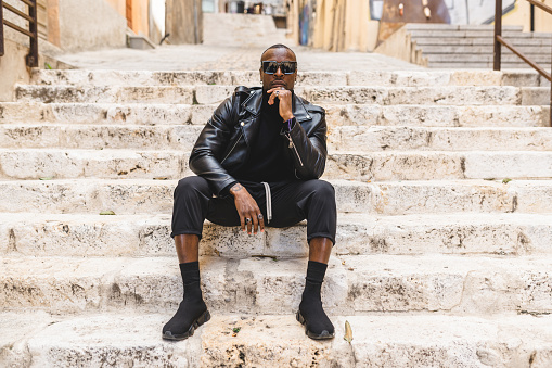 Full body brutal calm African American male in black outfit and leather jacket sitting on stone stairs in old city and looking at camera