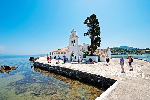 Tourists visiting the Holy Monastery of Panagia Vlacherna, Island of Corfu, Greece. The Vlacherna Monastery is a 17th-century Christian monastery on a small islet connected by a wooden pedestrian walkway to the mainland. Fishing and ferry boats run from thee islet to Pontikonisi Island (Mouse Island). The monastery is now only used for some celebrations but is open to the public. Corfu, or Korkyra, lies to the west of the Greek and Albanian mainland and separates the Adriatic from the Ionian Seas. Largely formed from Limestone, the island measuring some thirty miles by 18 miles is now a tourist destination, known for its beaches, climate and olives. Medieval castles and ancient ruins chart the military history of the island back to ancient Greek mythology.
