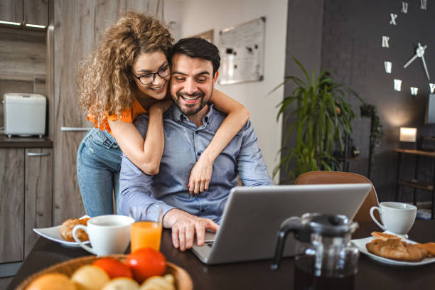 istock Lovely girlfriend hugging her boyfriend while he is working on laptop, eating breakfast and drinking coffee 1603117917