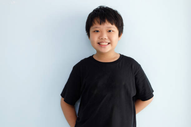 A cheerful Asian boy standing against a light blue wall, smiling, and looking at the camera