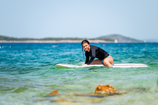 Young girl with black swimwear sitting on a white surfboard in the ocean on a sunny day in Primosten, Croatia.