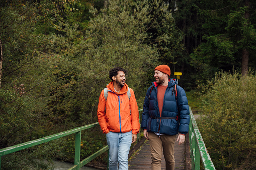 A  male same-sex couple walks across a bridge as they cross a river in a woodland area while hiking in Austria together. They are wearing casual autumn clothing.