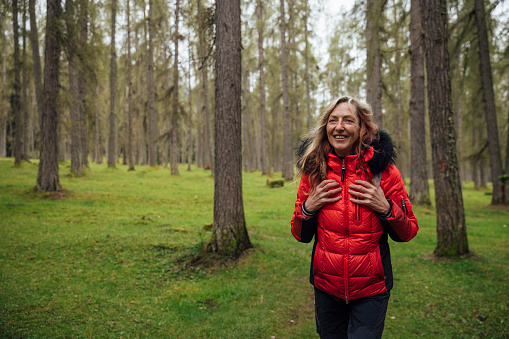 A woman hikes through an Austrian forest during Autumn. She is wearing a red down jacket and backpack. She is smiling and showing positive emotion as she walks through the woodland of Larch trees.