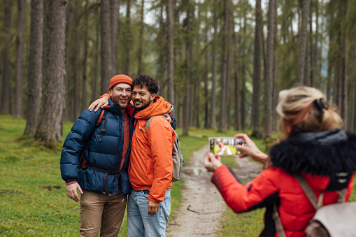 A female friend stops to take a photo on her smart phone of her friends who are a real gay couple. They pose as she holds up the phone as she stand near a dirt track while out hiking in a woodland of Larch trees in Austria.