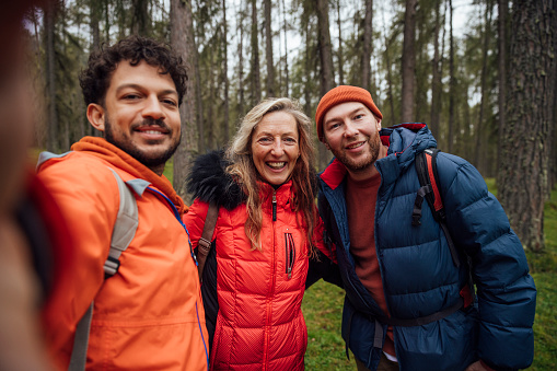 A male friend takes a photo selfie of his female friend and male partner who are a real gay couple. They pose as he holds out his arm and look into the camera's point of view while standing side by side close together while out hiking in a woodland of Larch trees in Austria. They are laughing.