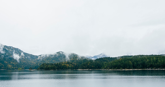 A landscape of Lake Eibsee in the German Bavarian Alps. The lake is calm and the water is glassy on a overcast day. The evaporation of moisture rises from between the trees in the distance.