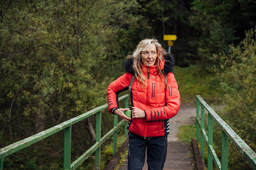 Radiating positivity, a mature woman in a red puffer jacket walks across a bridge. Retrieving her smartphone from her pocket, she readies to capture the serene before her. Behind she leaves wooded area of trees from where she came.