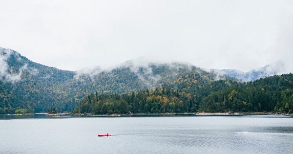 A landscape of fLake Eibsee in the German Bavarian Alps. The lake is calm as a kayaker paddles across the water on an overcast day. The evaporation of moisture rises from between the trees in the distance.