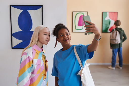 Waist up portrait of two teenage girls taking selfie photo in modern art gallery or museum, copy space