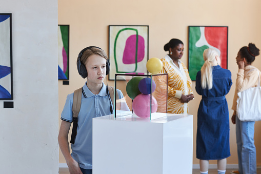 Portrait of schoolboy wearing headphones looking at abstract art sculpture in museum, copy space