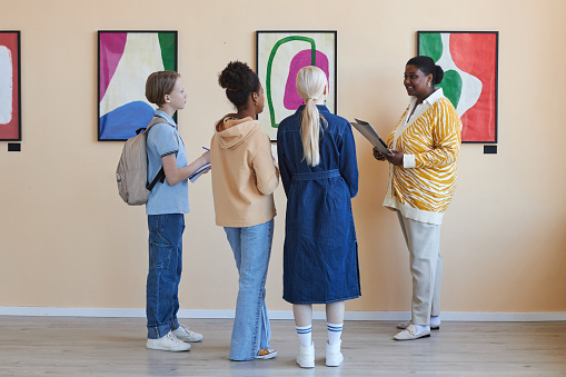 Colorful back view diverse group of teenagers listening to female teacher in modern art gallery