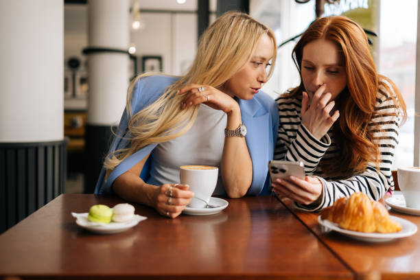 istock Front view of two happy charming girlfriends using mobile phone sitting at the cafe indoors and pointing finger. Beautiful best female friends laughing together 1603195347