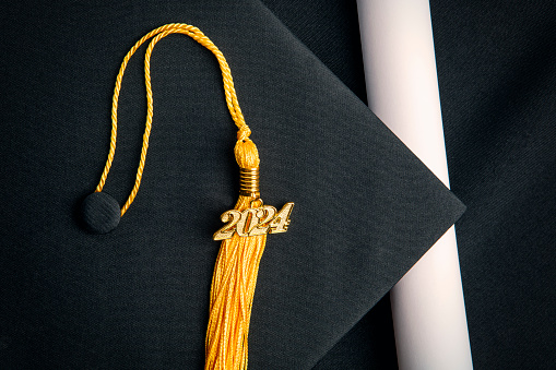 A class of 2024 charm on a yellow tassel on black graduation cap with a rolled diploma.