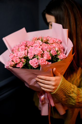 Young darkhair woman florist holding in hands large bouquet of fresh pink roses wrapped in pink paper and decorated with satin ribbon