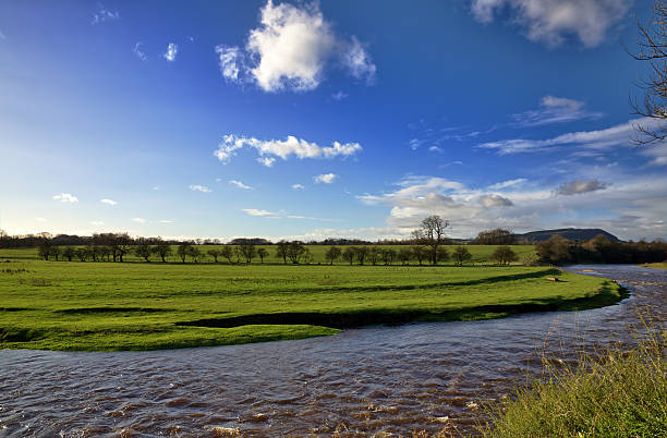 A peaceful view of the River Ribble, near Clitheroe, Lancashire, running through verdant green meadows, and set again a blue sky with clouds, on a sunny autumn day.