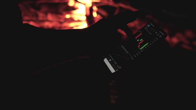 Businessman Using Mobile Phone For Checking Stock Chart At Night With Camping Bonfire In Background