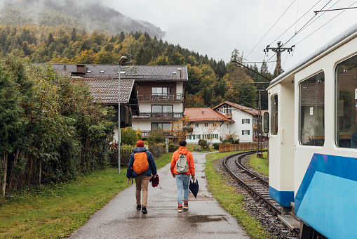 Full rear view shot of a mid-adult same-sex couple walking along a footpath next to a commuter train railway. They are both holding umbrellas. They are located in Garmisch, Germany.