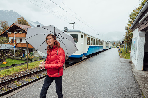 Three-quarter-length shot of a mature female walking along a train station platform next to a commuter train going past. She is wearing a vibrant red jacket and holding an umbrella. She is located in Garmisch, Germany.