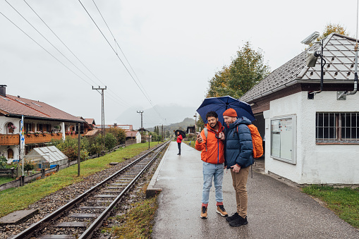 Full shot of same-sex couple waiting on a train station platform. They are both wearing warm clothing. One of the men is holding a phone and an umbrella. They are located in Garmisch, Germany.