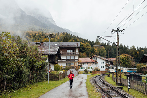 Full shot of a mature female walking along a footpath next to a commuter train railway. She is wearing a vibrant red jacket and holding an umbrella. She is located in Garmisch, Germany.