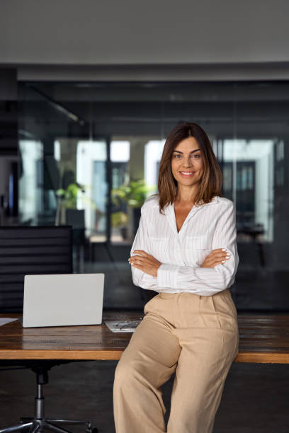 Vertical photo of Hispanic business woman with crossed arms smiling at camera. European or latin confident mature good looking middle age leader female businesswoman on office background, copy space. stock photo
