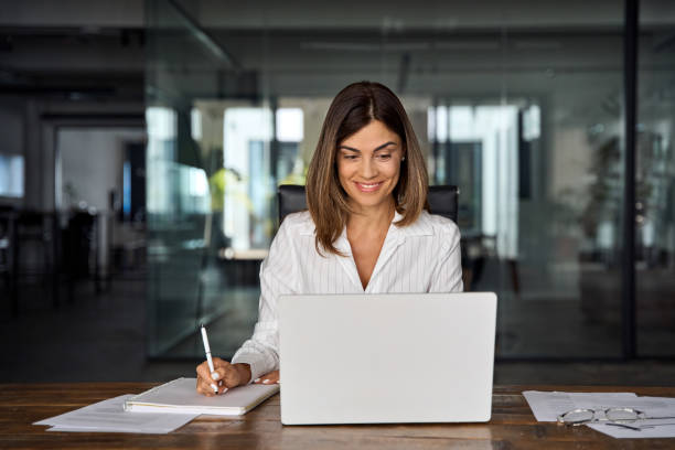 40s mid age European business woman CEO working on laptop sitting at table workspace, making notes in office. Smiling Latin Hispanic mature adult professional businesswoman using pc digital computer stock photo