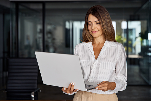 European businesswoman CEO holding laptop using fintech application standing at workplace in office. Smiling Latin Hispanic mature adult professional business woman using pc digital computer.
