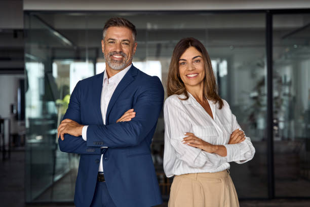 Portrait of smiling mature Latin or Indian business man and European business woman standing arms crossed in office. Two diverse colleagues, group team of confident professional business people. stock photo