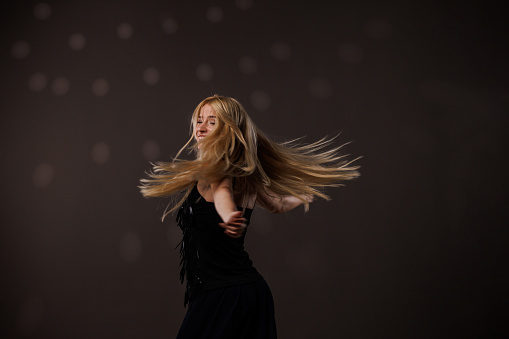 Portrait of cool young blond woman having fun and enjoying dancing against gray background with disco ball throwing reflection on her. Her hair flying all over due to dancing.