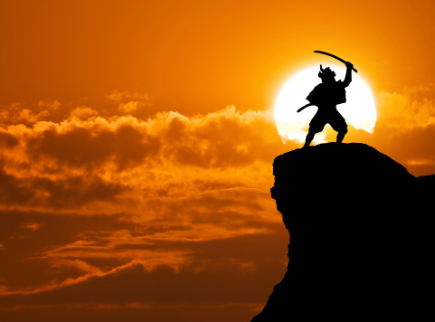 The silhouette of a samurai raises his Wakizashi to the sky while standing on top of a mountain.  Behind him, the sun sets with the sun directly behind the figure of the samurai.  The sky features a bright red and orange sunset with several clouds mingling around the sun.
