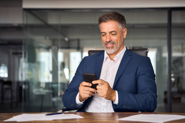 istock Middle aged Hispanic business manager ceo using cell phone mobile app, laptop. Smiling Latin or Indian mature man businessman holding smartphone sit in office working online on gadget with copy space. 1603245536