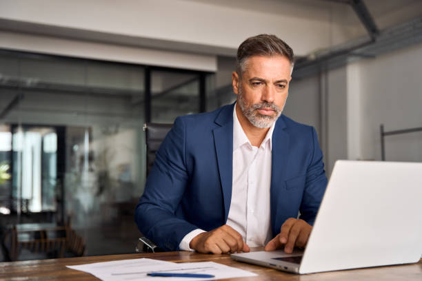 istock Mature Indian or Latin business man ceo trader using computer, typing, working in modern office, doing online data market analysis, thinking planning tech strategy looking at laptop with copy space. 1603245542