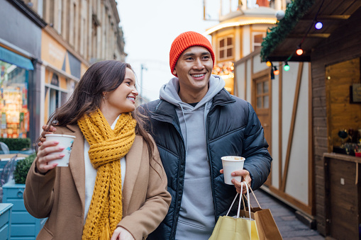 City sidewalks and warm cocoa in hand, a couple's laughter mingles with the festive buzz as they Christmas shop, creating a scene of shared joy amidst the holiday lights.