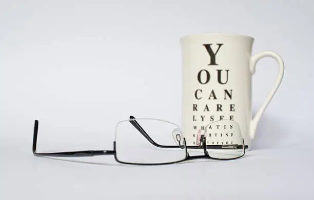 A pair of eye glasses in front of a mug with funny eye chart. Mug is blurry except the portion where the eye glass lens is.