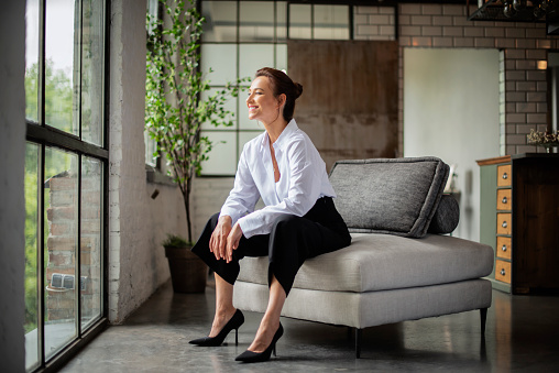 Portrait of beautiful mid aged woman relaxing in an armchair by the window. Attractive female wearing white shirt and black pants. Full length shot.