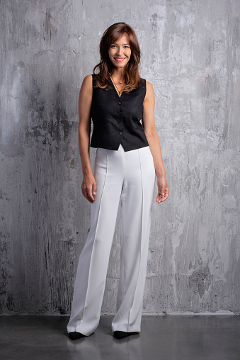 Full length of a beautiful brown-haired woman standing in front of a grey wall. Cheerful smiling female wearing black shirt and white pants and looking at camera. Copy space.