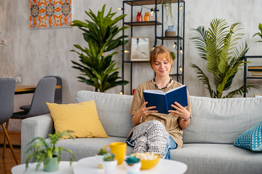 Smiling young woman sitting on the sofa and reading a book at home.