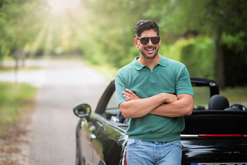 A handsome middle-aged man standing next to his convertible car and laughing. Confident man wearing sunglasses and casual clothesl