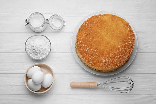 Plate with delicious sponge cake and ingredients on white wooden table, flat lay