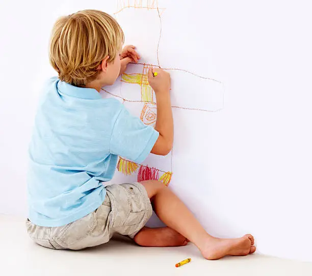 A rear view shot of a young boy drawing a picture on a wall with crayons