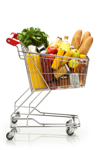 Side view of a metal shopping cart filled with a large variety of colorful groceries that includes some fresh vegetables, fruits, canned food, fruit juice, cooking oil and three loafs of bread. The cart is standing on reflective white background producing a soft reflection under it. The cart has a red plastic handle. DSRL studio shot with Canon EOS 5D Mk II  