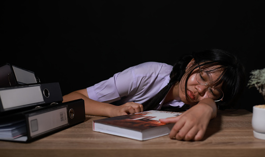 Tired young schoolgirl in student uniform with eyeglass sleeping sitting at an uncomfortable work desk in the room. Exhausted girl student resting after a long read book at night.