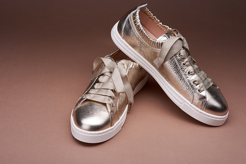 Fashionable golden metallic sneakers with wrinkled leather details and white rubber soles on a gradient brown background. Close-up view. The concept of modern stylish footwear